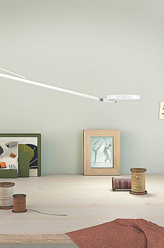 Bureau en bois clair avec lampe de bureau blanche, cadres photos et bobines de fil. Tissu plié sur la table.