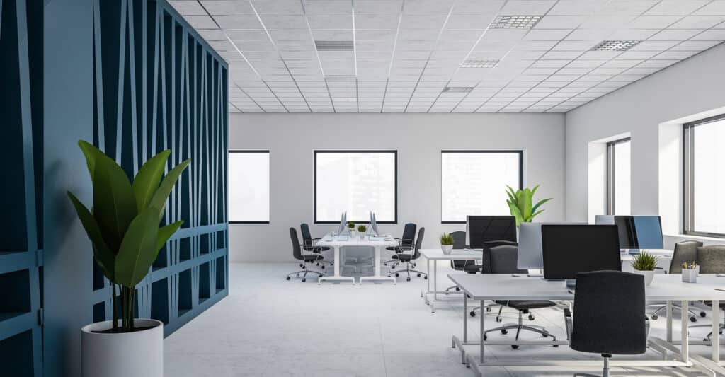 Open space de bureau moderne et lumineux avec cloisons bleues, bureaux blancs, ordinateurs, chaises noires et plantes, grandes fenêtres.