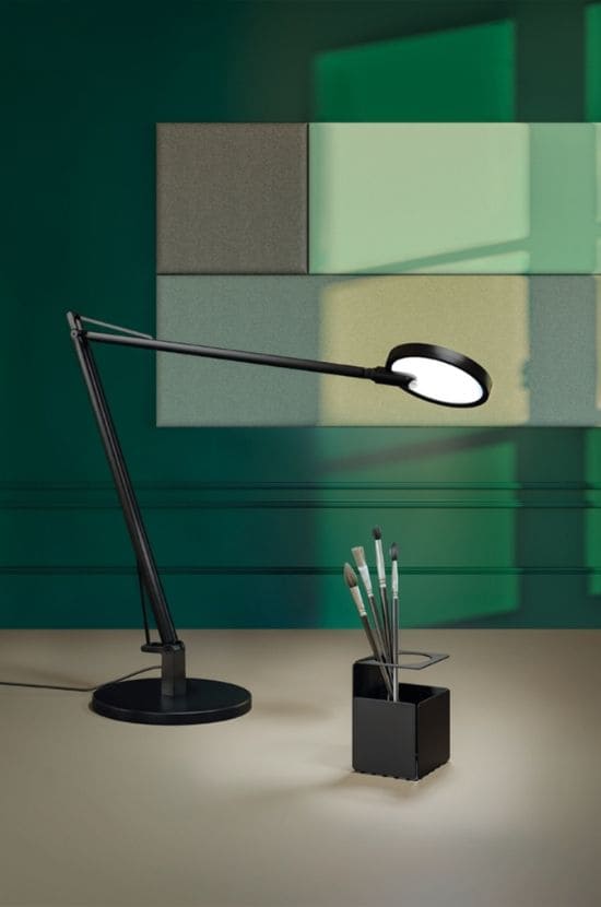 Lampe de bureau noire articulée avec tête lumineuse blanche et pot de pinceaux sur bureau clair. Mur vert, panneaux acoustiques.