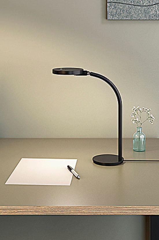 Lampe de bureau LED noire moderne sur un bureau clair, éclairant une feuille de papier avec un stylo. Un vase de fleurs ajoute une touche décorative.