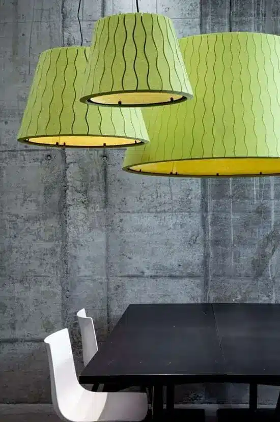 Trois lampes suspendues vert clair à motifs ondulés au-dessus d'une table noire, devant un mur en béton gris. Deux chaises blanches visibles.