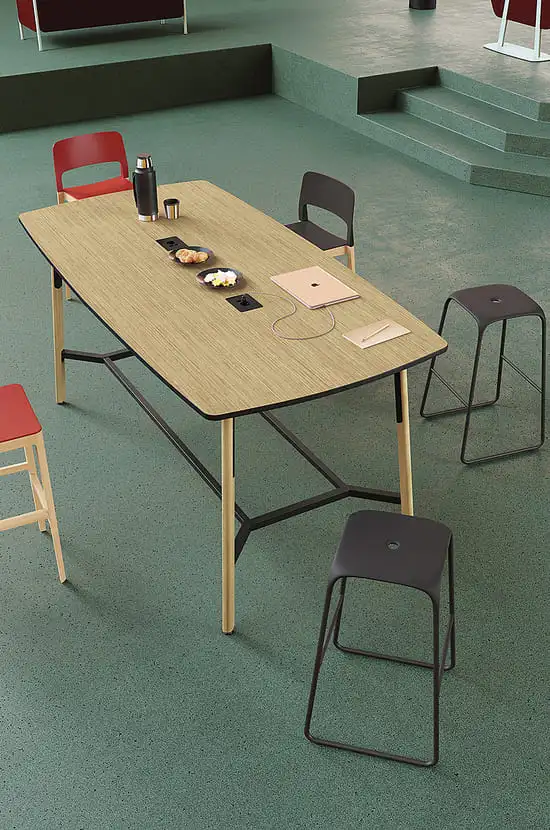Longue table de réunion bois clair avec laptop, prises et snacks. Chaises et tabourets rouges/noirs dans un espace de bureau moderne vert.