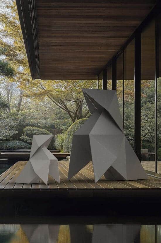 Deux sculptures géométriques grises, style origami, sur une terrasse en bois. Jardin luxuriant et étang avec reflets, fond de bâtiment moderne.