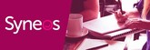 Logo Syneos magenta. Deux personnes collaborent à une table, une main écrit sur un carnet près d'une tasse de café, suggérant une réunion d'affaires.